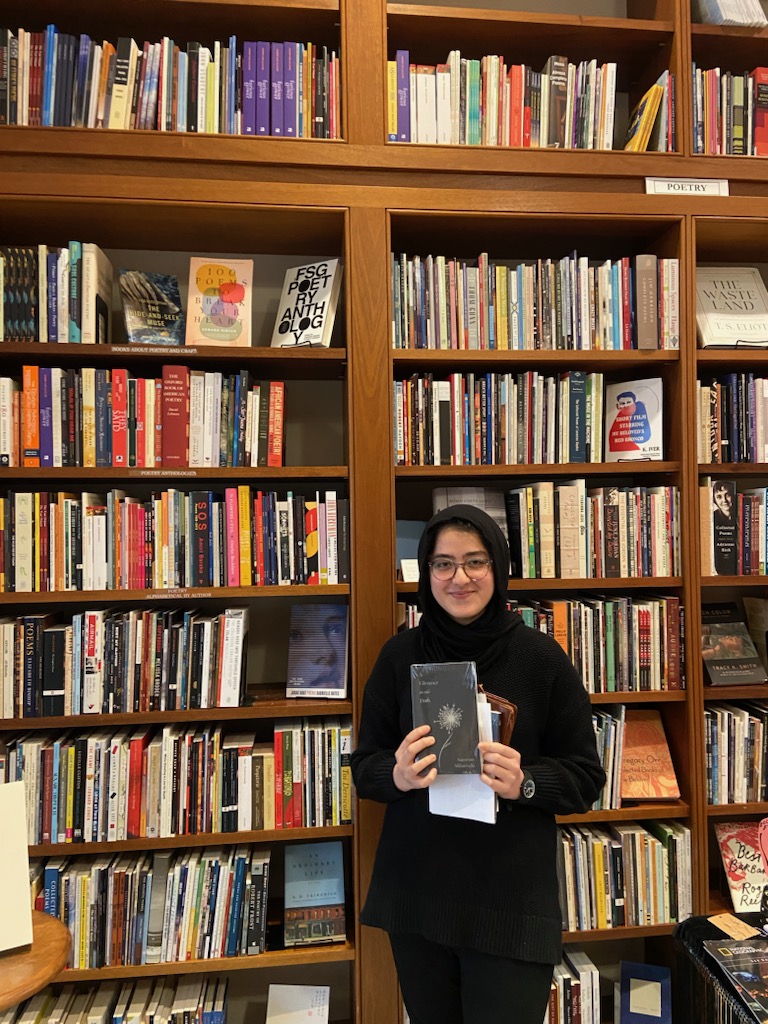 Akbarzada stands in front of a tall bookcase holding her book A Glimmer in the Dark