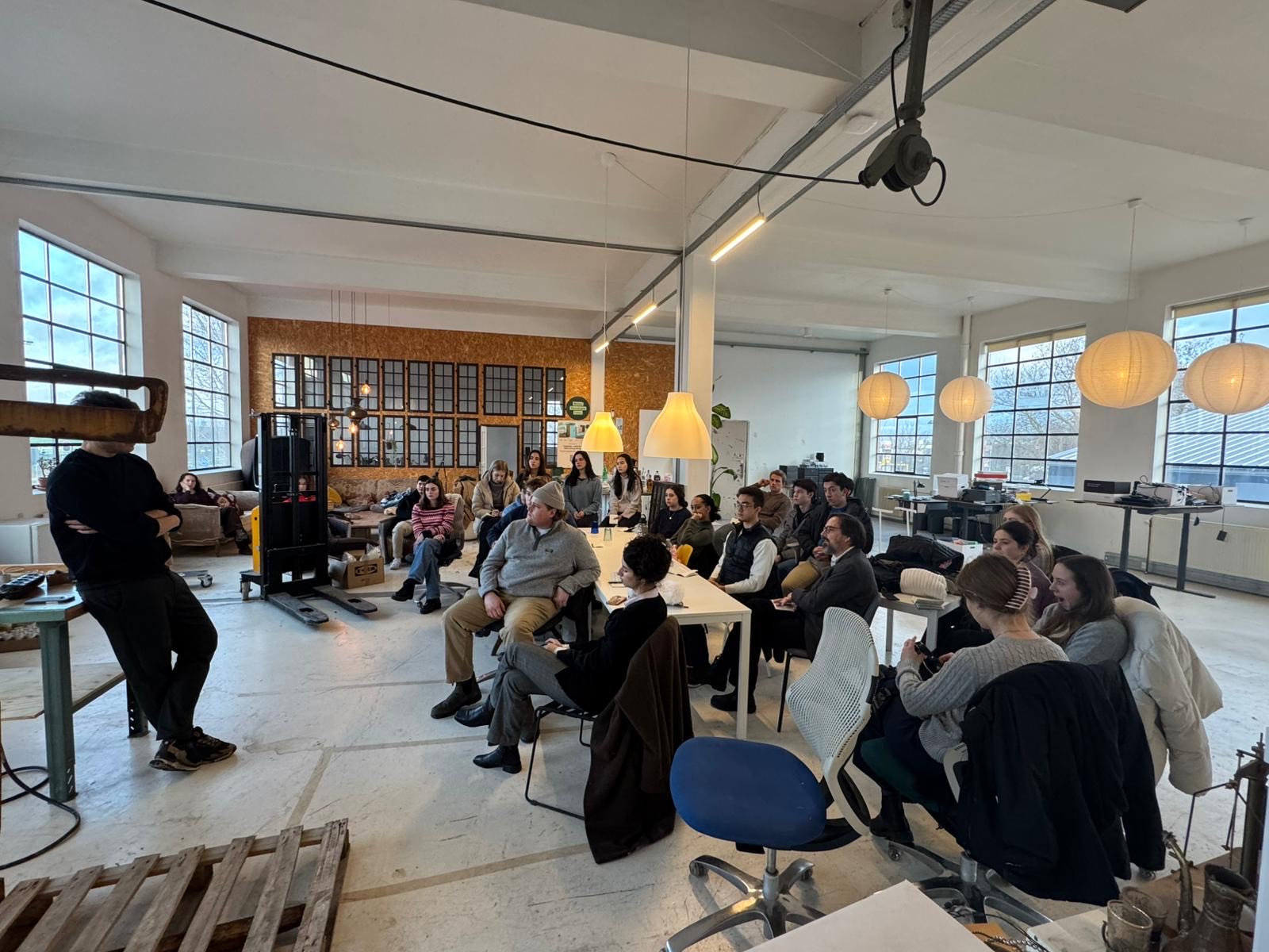 Students sit in an industrial room, one standing person speaks to the class
